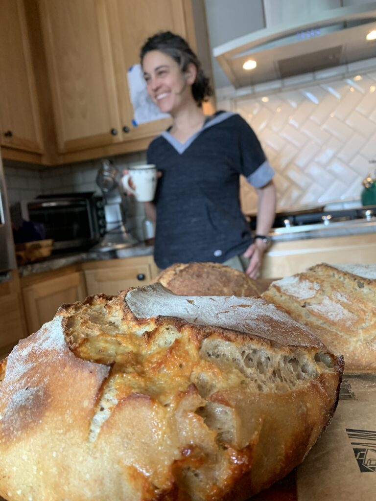 Kera smiles in the background as two fresh loaves of sourdough bread cool on the counter in the foreground.
