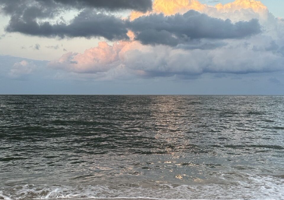 Storm clouds part over the ocean at dusk, their edges lit gold and pink, with the light reflecting on the water below.